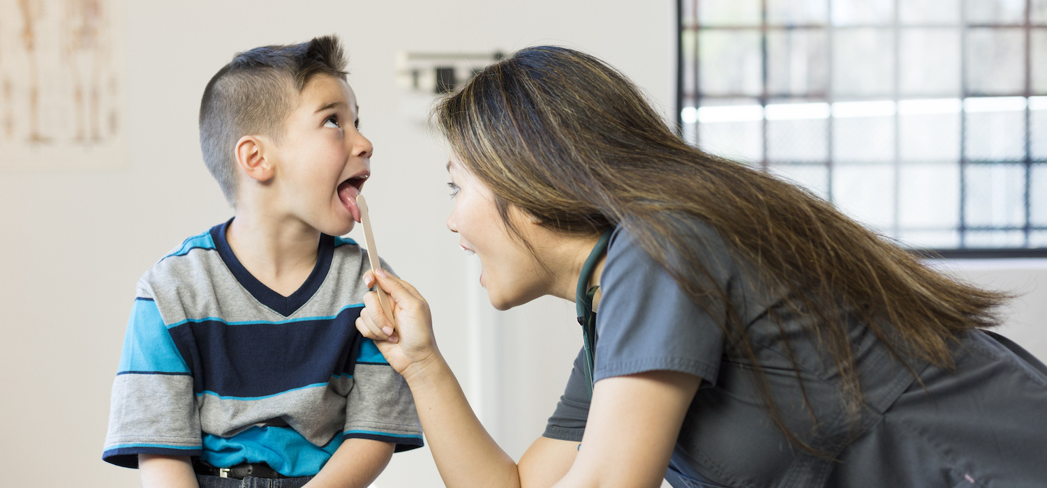 An asian doctor examines a young hispanic boy in a doctor's office. The nurse wears dark blue scrubs and uses a tongue depressor while she examines the boy's throat. The boy sits on an examination table with his mouth open.