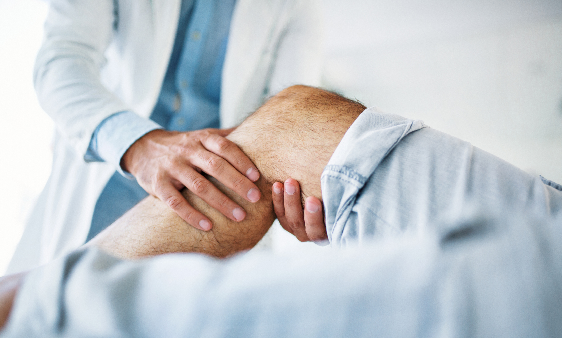 Closeup side view of early 30's unrecognizable doctor examining a knee of a senior gentleman during an appointment. The doctor is gently touching the tendons around the knee and the knee cap and trying to determine the cause of pain.